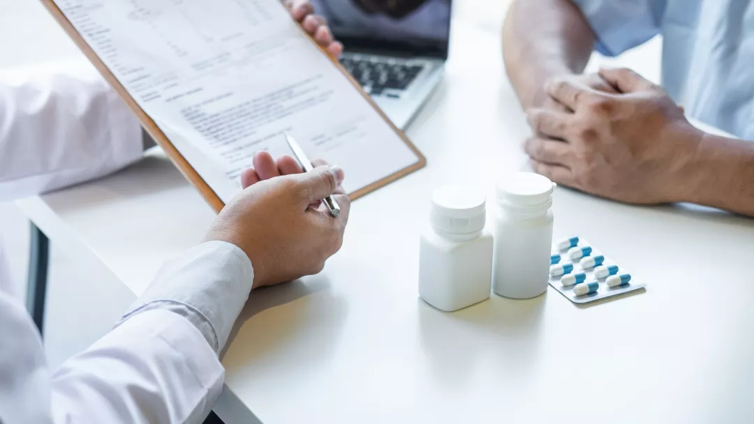 A doctor reviews a prescription with a patient, discussing medication details and health records at a desk. The image highlights the importance of thorough medical consultations and detailed patient care.