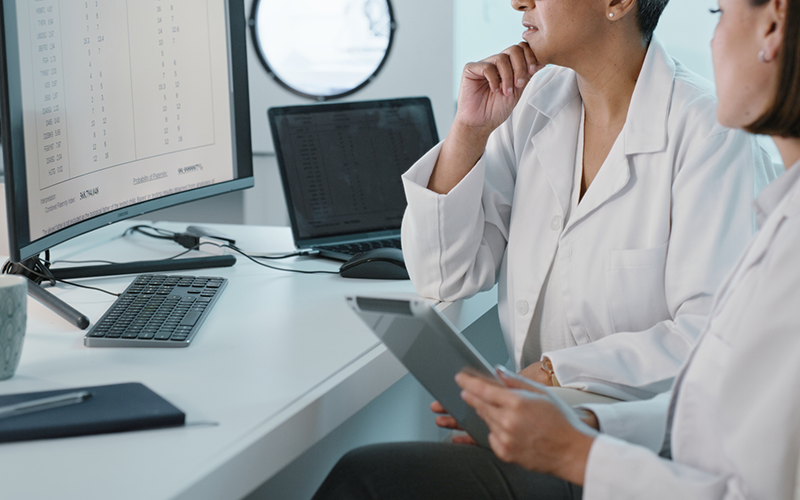 Profissionais de saúde analisando dados clínicos em estações digitais Dois profissionais de jaleco observam um monitor com tabelas enquanto consultam um tablet, em uma mesa com teclado, laptop e caneca, sugerindo revisão de resultados ou prontuários eletrônicos.