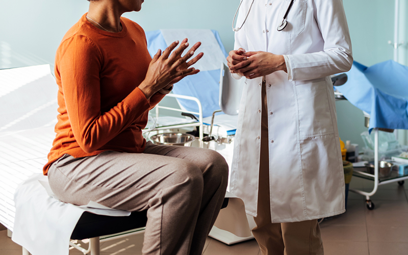 Paciente sentada conversando com profissional de saúde de jaleco em um consultório, com equipamentos ao fundo
