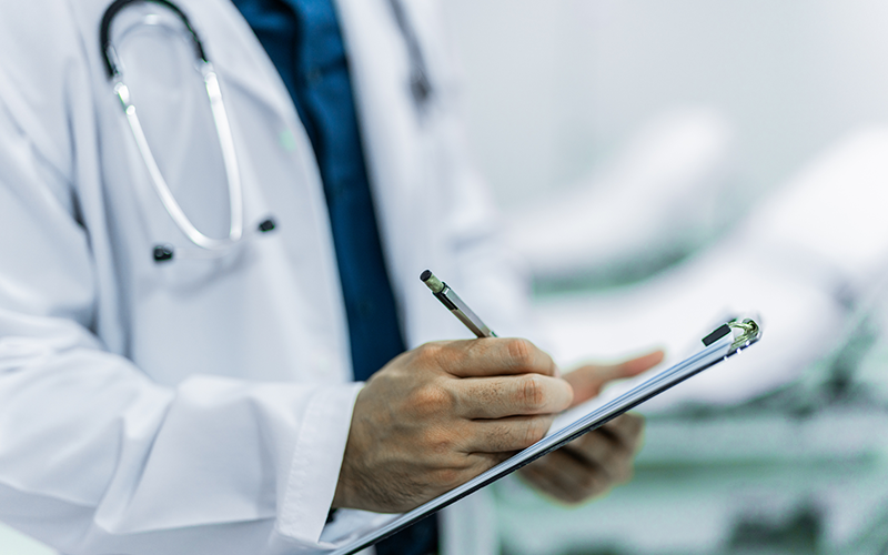 Profissional de saúde em jaleco escreve em prancheta em ambiente clínico.