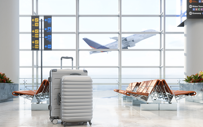 Mala de viagem prateada em saguão de aeroporto com aeronave decolando ao fundo