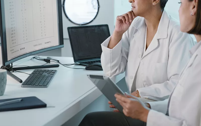 Profissionais de saúde analisando dados clínicos em estações digitais Dois profissionais de jaleco observam um monitor com tabelas enquanto consultam um tablet, em uma mesa com teclado, laptop e caneca, sugerindo revisão de resultados ou prontuários eletrônicos.