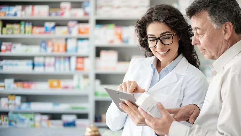 Dois profissionais em ambiente farmacêutico discutindo informações sobre um produto, com prateleiras de medicamentos ao fundo.