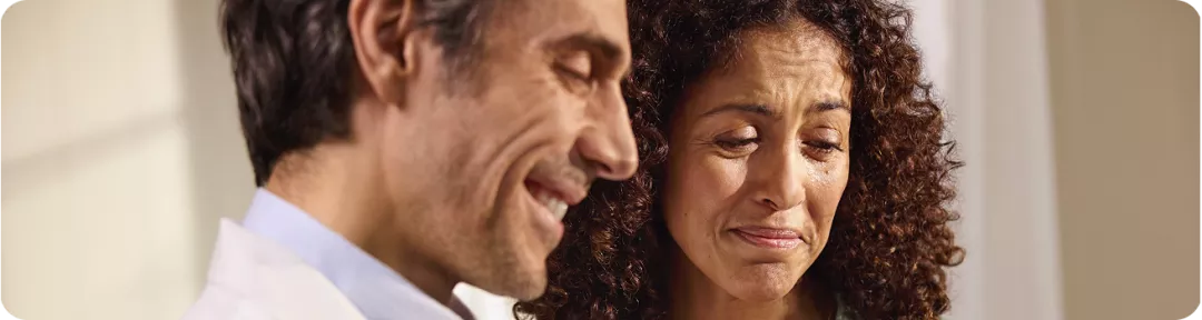 Close de um profissional de saúde usando jaleco branco e camisa azul claro, em pé ao lado de uma paciente em um ambiente interno bem iluminado, com paredes claras ao fundo, sugerindo uma situação de atendimento ou acolhimento em consultório.