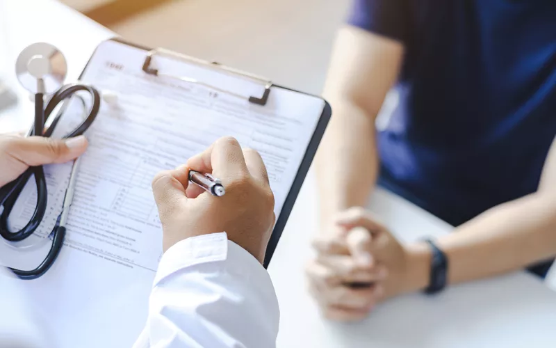 Profissional de saúde preenche formulário em prancheta durante consulta; paciente à frente com mãos entrelaçadas e estetoscópio visível.