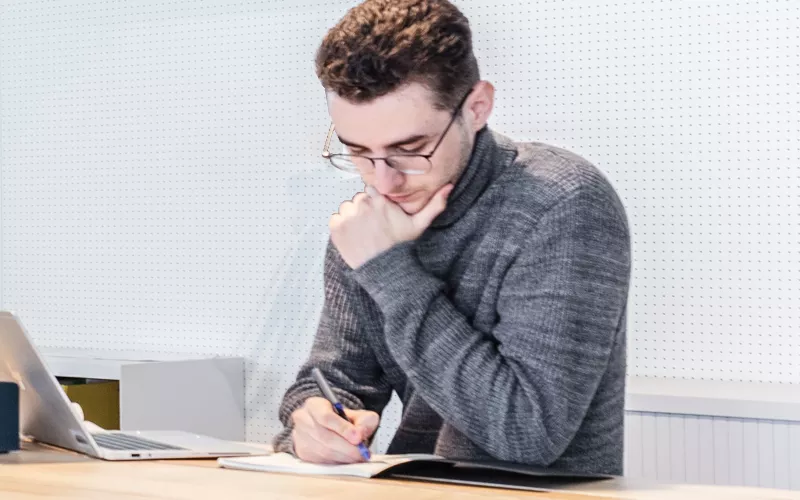 Pessoa sentada em mesa de trabalho, escrevendo em caderno com caneta ao lado de um laptop em ambiente de escritório claro