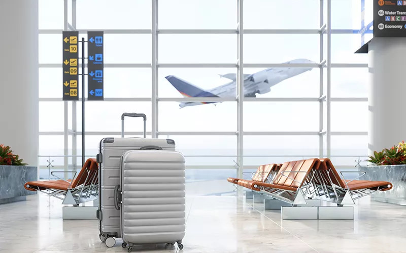 Mala de viagem prateada em saguão de aeroporto com aeronave decolando ao fundo